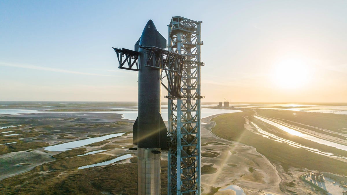 SpaceX's Starship rocket at a launch gantry, with the sun low on the horizon behind it.