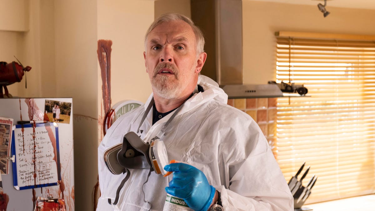 A large, bearded crime scene cleaner in a white hazmat suit poses with his cleaning spray