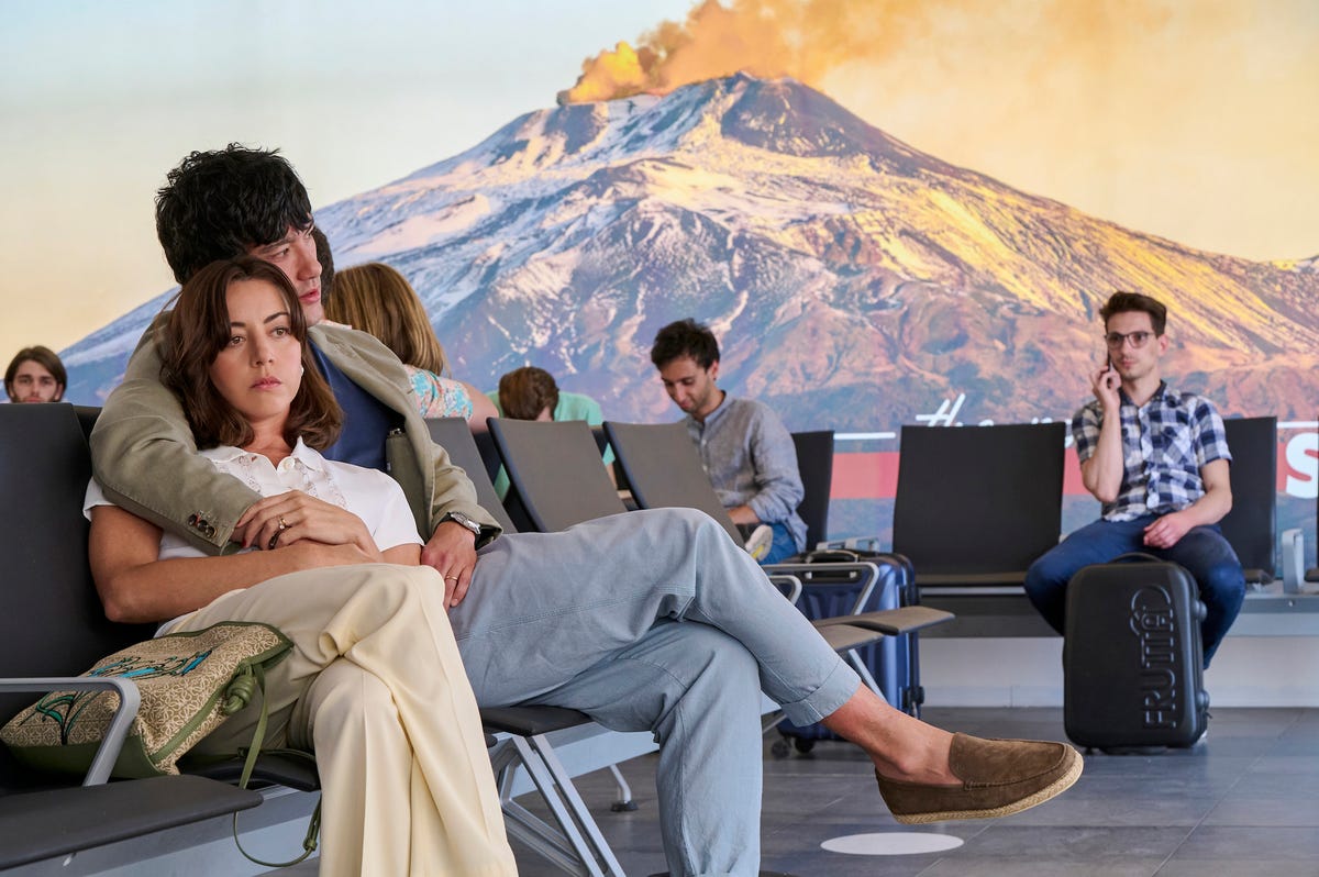 Aubrey Plaza and Will Sharpe as Harper and Ethan, sitting in each other's arms in an airport with an erupting volcano in the background