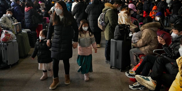 A woman and children wearing face masks walk by masked travelers wait at a departure hall to catch their trains at the West Railway Station in Beijing, Sunday, Jan. 15, 2023.