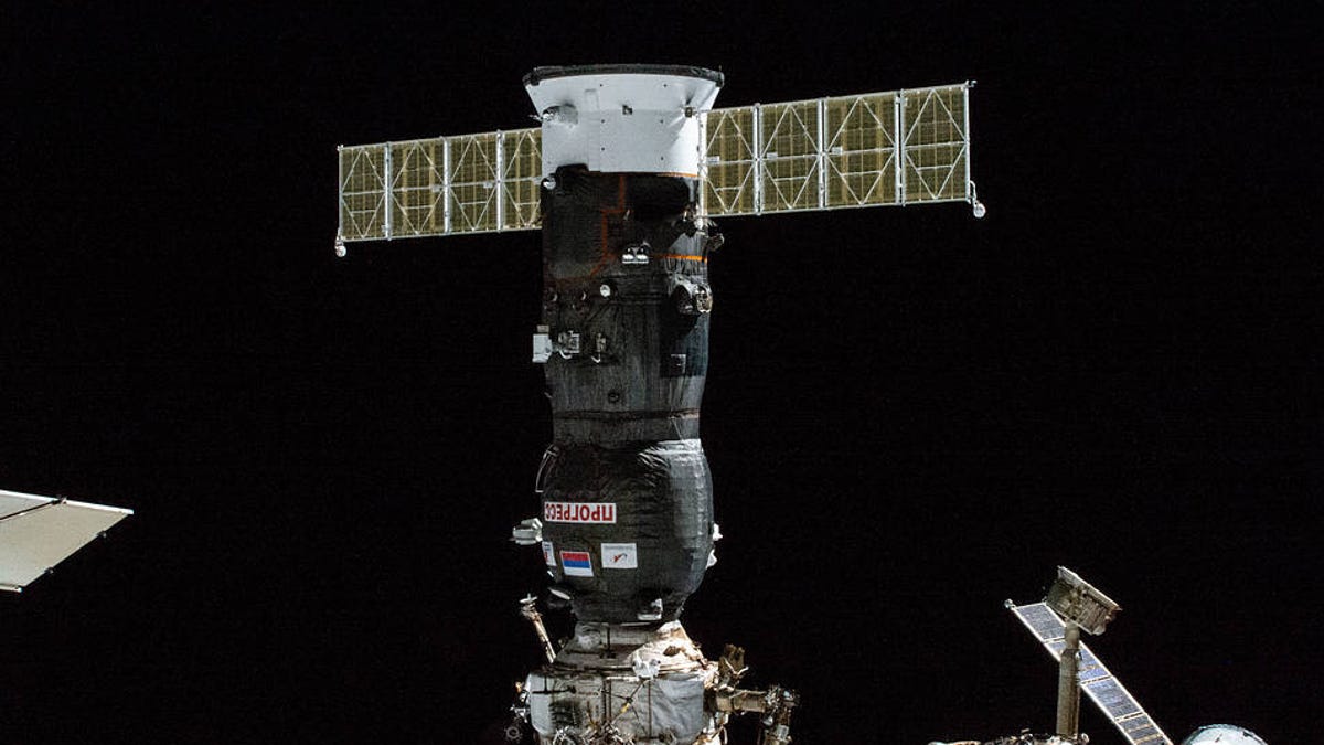 Russian Progress 82 cargo craft, a dark cylinder with outstretched solar panels like a cross, is connected to a dock on the ISS.