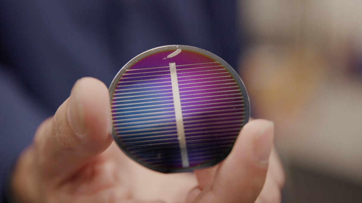 A hand holds a round, purplish solar cell with small lines running across it. It's a prototype made from lunar regolith simulant.