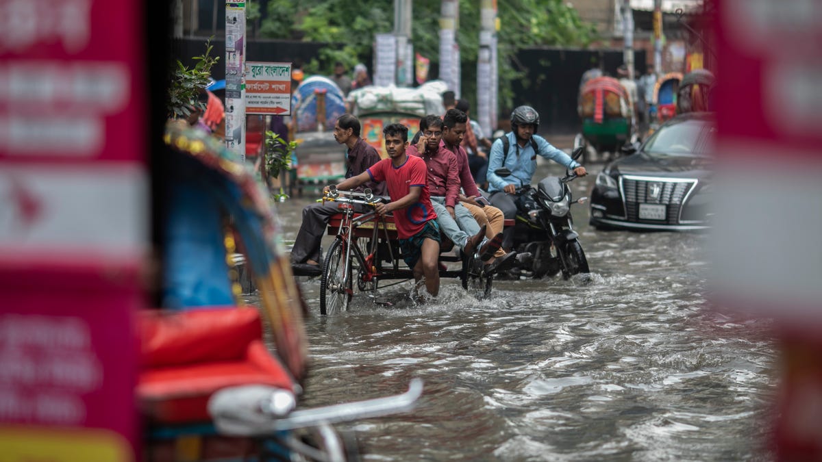 In a flooded roadway, men push or ride a rickshaw and a bike through knee-deep water, followed by a car.