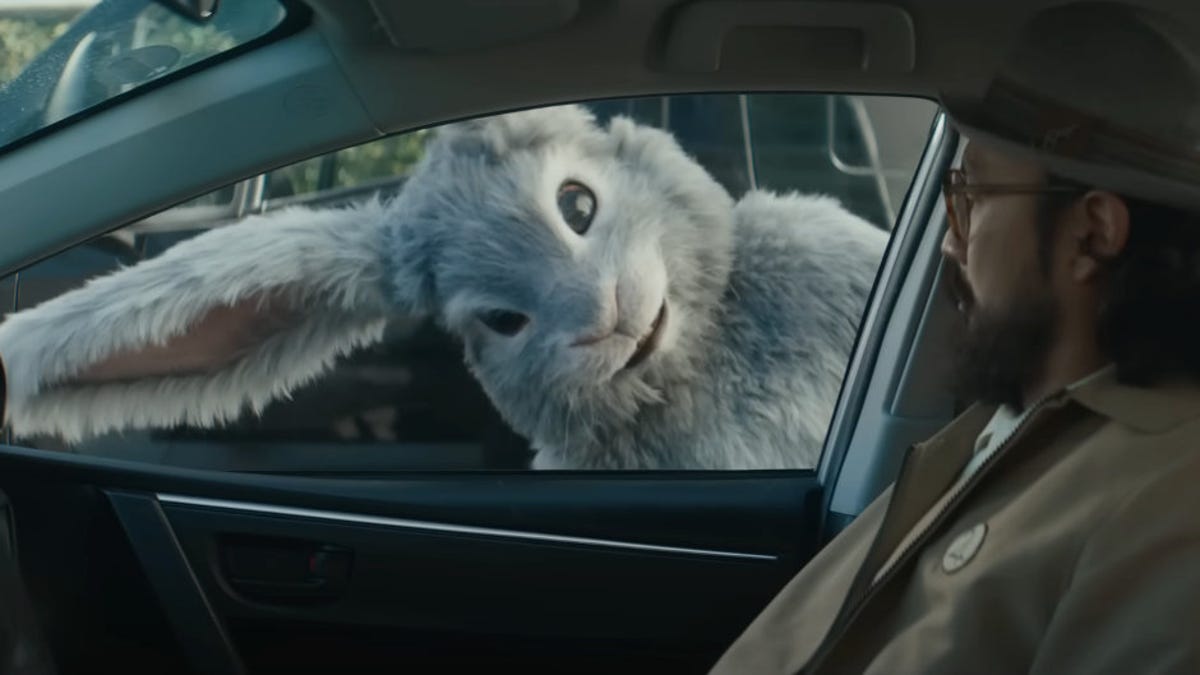 Tubi Super Bowl ad still shows a large gray rabbit leaning over and peering into a car window as a man in a khaki jacket and hat stares back.