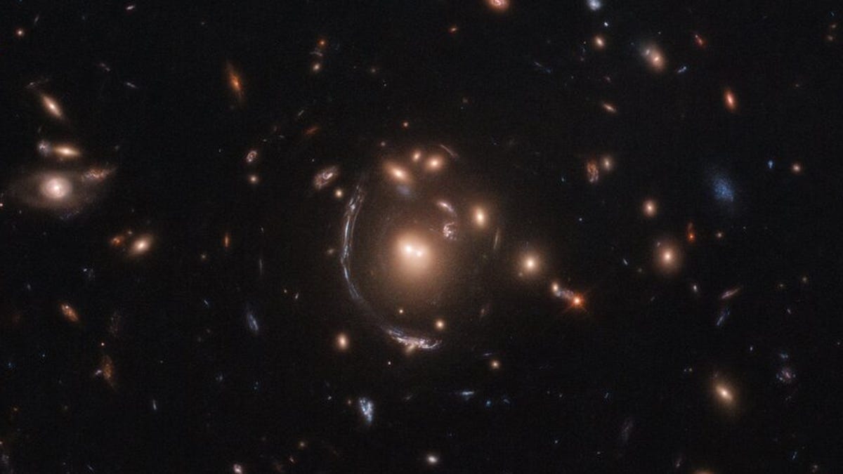 A dark background is scattered with hazy, glowing galaxies. At the center, one galaxy appears stretched out as a long arc instead of having a characteristic spiral, for instance.