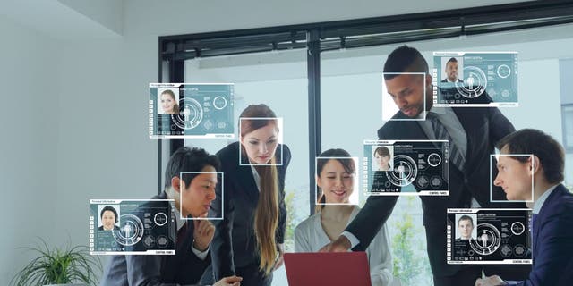 A group of co-workers gather around a table and use facial recognition while looking at a laptop. 