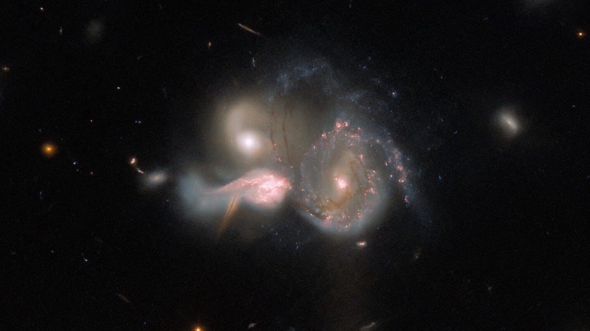 Three galaxies. They are close enough that they appear to be merging into one. Their shapes are distorted, with strands of gas and dust running between them. Each is emitting a lot of light. The background is dark, with a few smaller, dim and faint galaxi