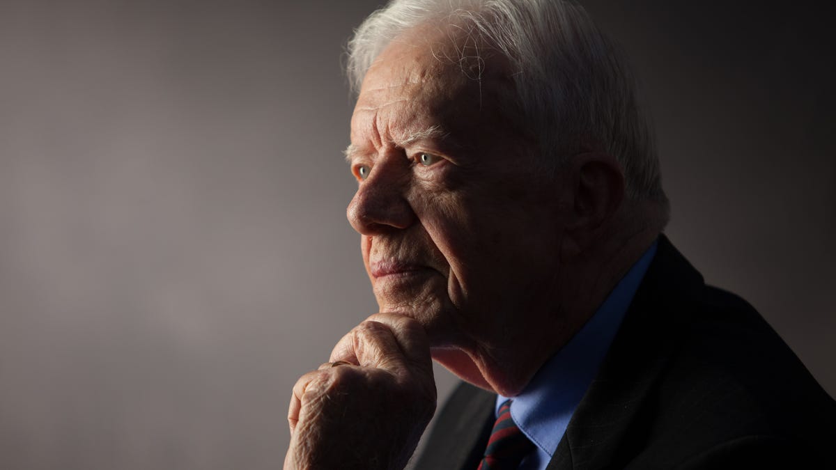 Carter in three-quarters profile, looks off thoughtfully to the left, hand on chin, with a slight smile on his face.