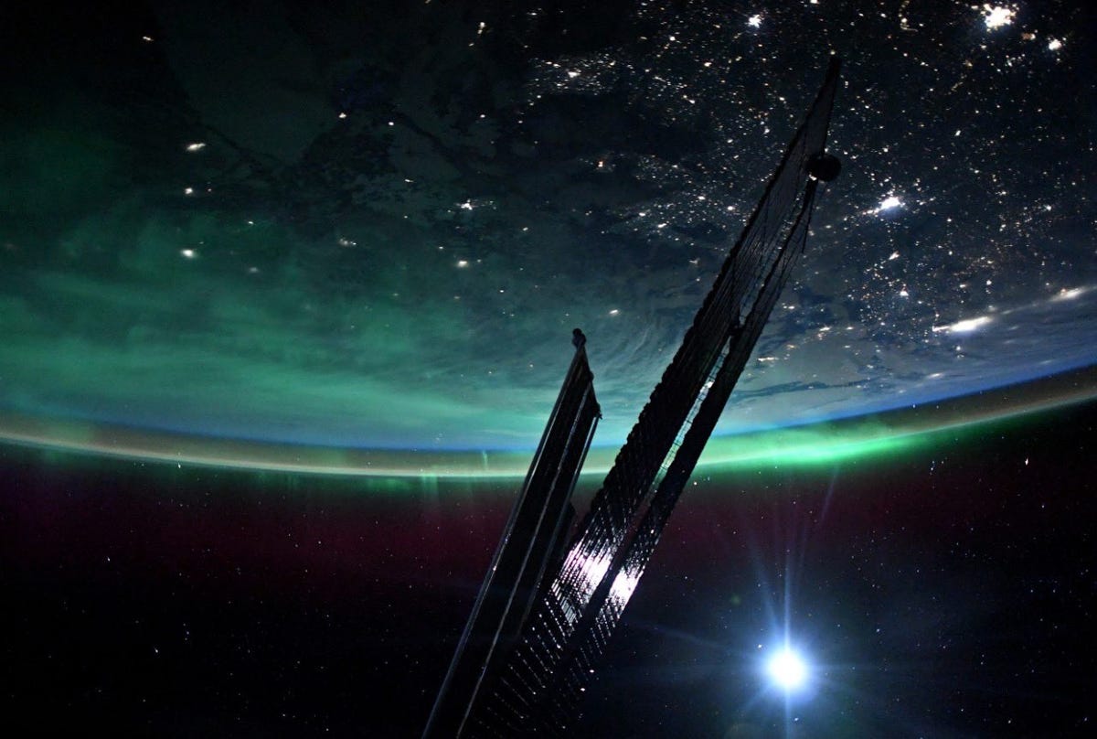 Photo of Earth taken from the ISS shows slim, dark shiny solar panels stretched across dark space lit by a lens flare. Part of Earth is visible upsdide-down with city lights below and green ethereal aurora along the curve.