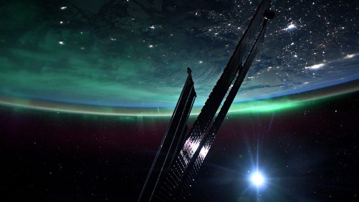 Photo of Earth taken from the ISS shows slim, dark shiny solar panels stretched across dark space lit by a lens flare. Part of Earth is visible upsdide-down with city lights below and green ethereal aurora along the curve.