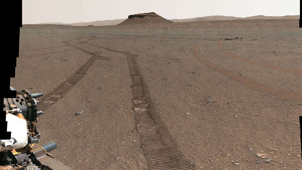 A vast landscape shows 10 very small, titanium sample tubes carefully placed along the surface of Mars by the Perseverance rover. The color of the landscape is a brownish-red and rover tracks mark up parts of the surface. The rover's robotic arm is shown along the bottom-left part of the image. Far off into the distant horizon, a rocky hill can be seen.