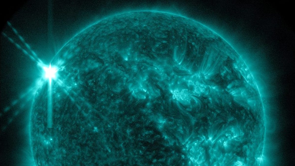 A teal Sun on a black background. The Sun has bright teal areas scattered across the star. On the top left of the star, there is a white X where the flare burst from the Sun.