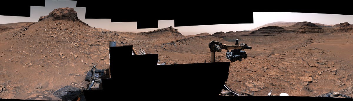Mars Gale Crater landscape panorama stitched together from Curiosity rover images shows part of the rover hardware in the middle and rocky outcrops and rippled rocks on the ground ahead.