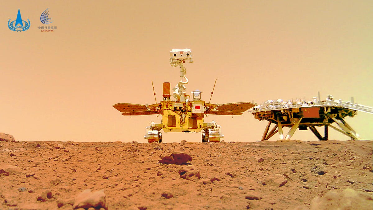 China's cute Zhurong rover "looks" at the camera with mast-mounted head and solar panels extending to the side. A leggy lander is visible in the background. Both are on a rocky, reddish Martian landscape.