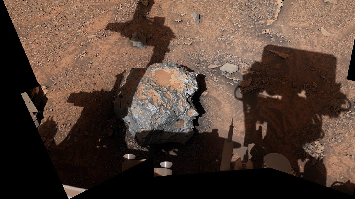 Color image of Mars ground with Curiosity rover throwing shadows over it. There's a shiny dark-gray iron-nickel meteorite on the ground with indentations.