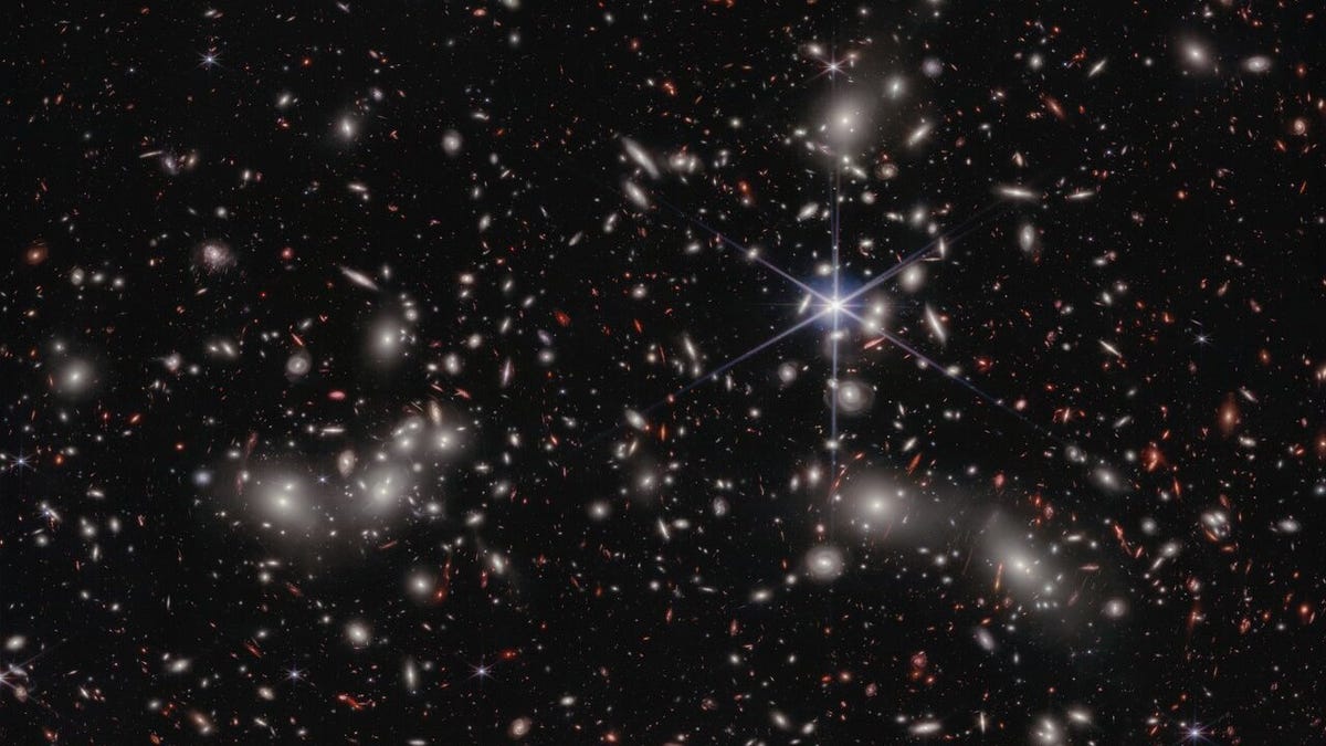 A crowded galaxy field on a black background, with one large star dominating the image just right of center. Three areas are concentrated with larger white hazy blobs on the left, lower right, and upper right above the single star. Scattered between these areas are many smaller sources of light; some also have a hazy white glow, while many other are red or orange.