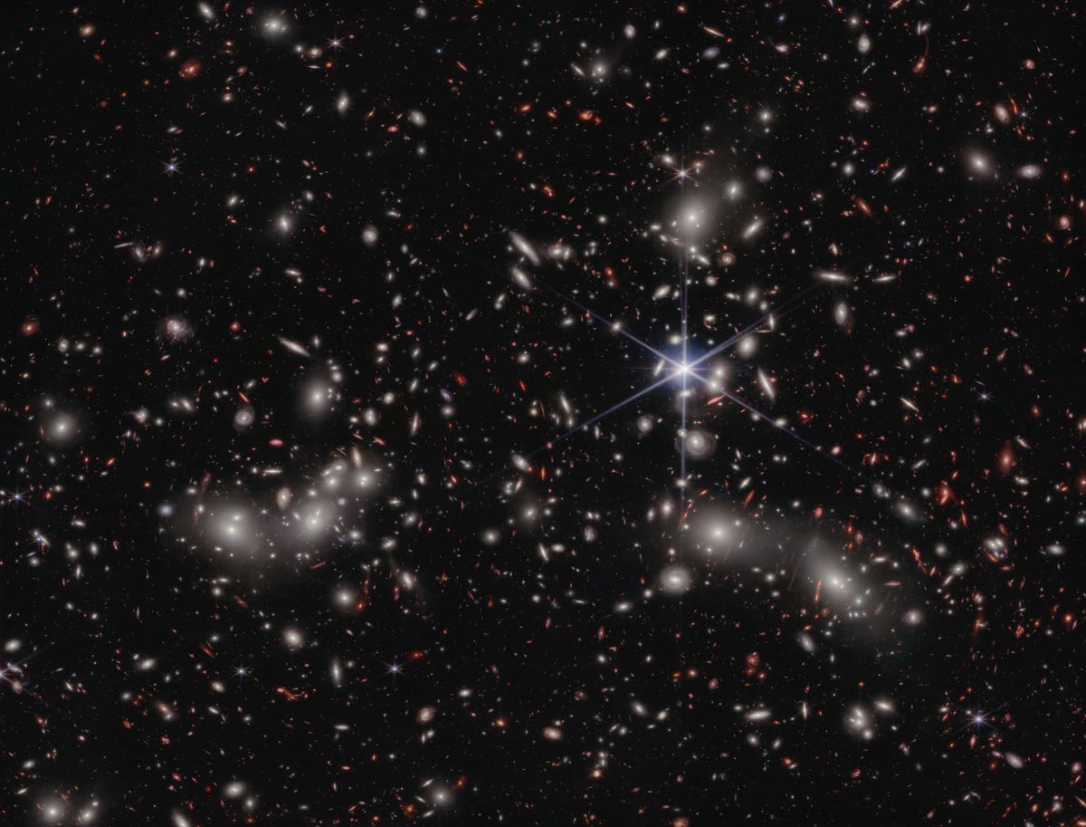 A crowded galaxy field on a black background, with one large star dominating the image just right of center. Three areas are concentrated with larger white hazy blobs on the left, lower right, and upper right above the single star. Scattered between these areas are many smaller sources of light; some also have a hazy white glow, while many other are red or orange.