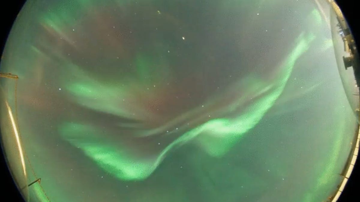 Cropped view of Swedish all-sky camera looking from horizon to horizon showing a dramatic ribbon of green, waving aurora light with hints of fuzzy red.