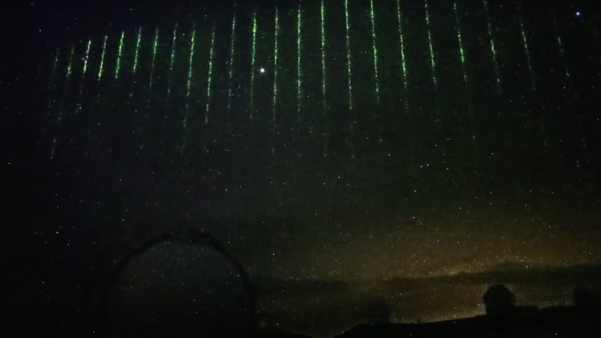 A still image captured by a camera on the Subaru Telescope in Hawaii shows the cloudy night sky with a curtain of individual downward streaks of green laser light.