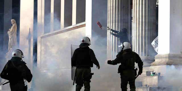 A hooded protester with a tear gas mask throws a fire extinguisher at riot police during clashes at the Athens University headquarters, in Greece, on March 8, 2023. Protests have broken out over the deadly train crash in Greece that killed 57 people. Three more people were charged in connection with the deadly train crash.