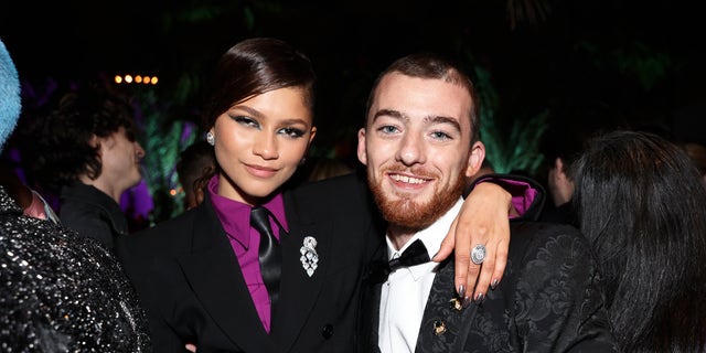 Zendaya in a purple shirt and black blazer with a tie puts her hand around a smiling Angus Cloud in a black tuxedo