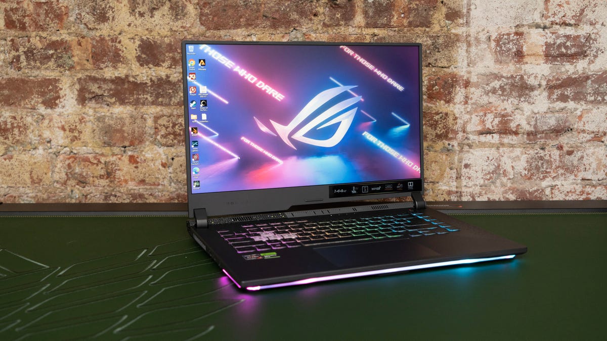The ROG Strix G15 open and angled to your right, with the RGB illumination on the keyboard and underglow, default ROG wallpaper on a pine green surface against a faded brick wall