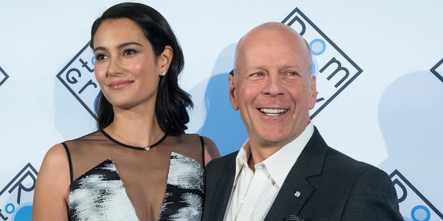 Emma Heming Willis looks to her right in a blue dress with black mesh as Bruce Willis looks to his left wearing a dark suit on the carpet