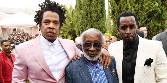 Jay-Z in a pink suit puts his arm around Clarence Avant and Diddy puts in a white suit and black shirt puts his arm around Clarence on the other side