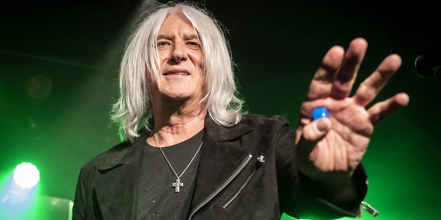 Joe Elliot reaches for the crowd in a black jacket with a cross necklace hanging around his neck