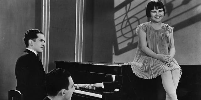 Baby Rose Marie sitting on top of a piano as she sings in a ruffled dress