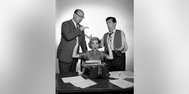 Rose Marie throwing her hands up in the air in front of a typewriter surrounded by papers as two guys argue