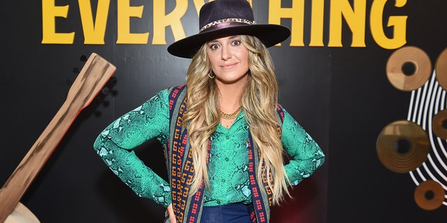 Lainey WIlson puts her hands on her hips in a vibrant green shirt and black rimmed hat ahead of the Academy Of Country Music Awards