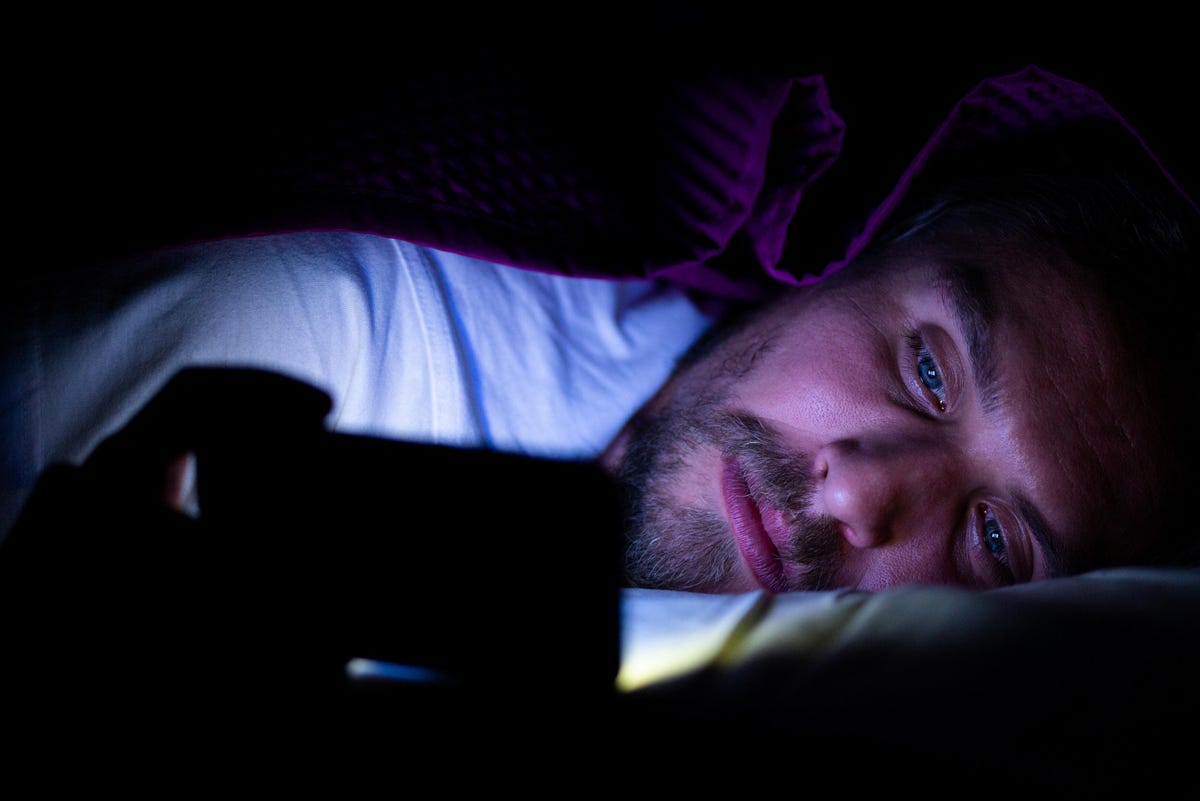 A man lays on a bed in a dark room, his face illuminated by the phone in front of him.