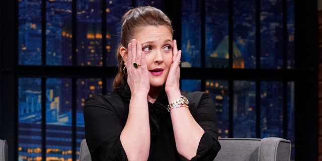 Drew Barrymore in a black dress puts her hands up to her face in distress on "Late Night with Seth Meyers"