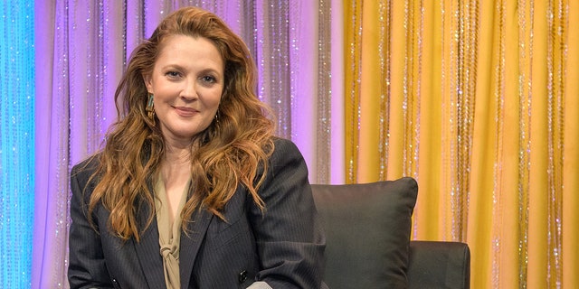 Drew Barrymore in a dark grey blazer with rolled up sleeves sits on a chair and soft smiles for the camera
