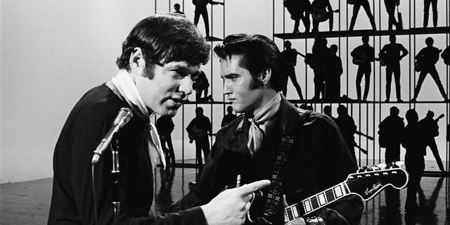 Steve Binder pointing in front of a camera as Elvis Presley looks away holding a guitar