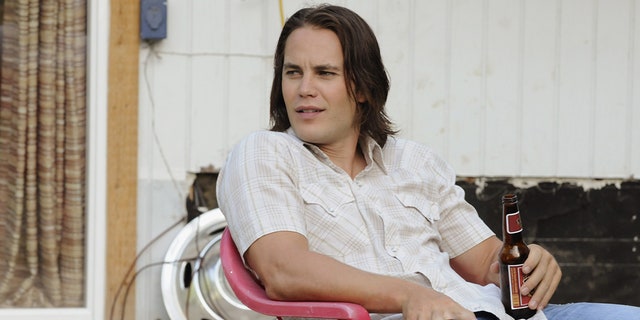 Taylor Kitsch as Tim Riggins in "Friday Night Lights" sits on a red chair in a white shirt and blue jeans