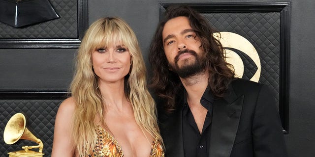 Heidi Klum in a gold low plunging sequin dress poses with husband Tom Kaulitz in a black suit and shirt