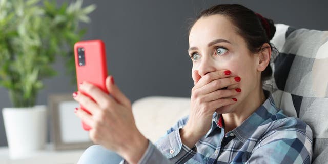 Woman covering mouth with hand, eyes wide in shock while holding up iPhone