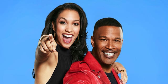 Jamie Foxx in a red leather jacket is hugged from behind by daughter Corinne pointing at camera