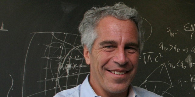 A close-up of Jeffrey Epstein smiling in a light blue collared shirt standing behind a blackboard