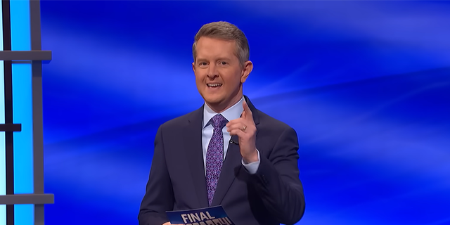 Ken Jennings in a navy suit with a light blue shirt and purple suit on the set of 'Jeopardy!' pointing at the camera