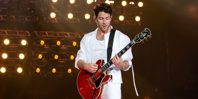 Nick Jonas in all white plays the guitar on stage and looks down at Yankee Stadium