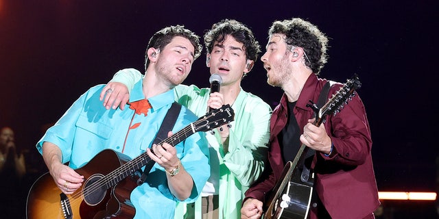 Nick Jonas playing the guitar leans back into Joe Jonas singing with Kevin Jonas, also playing the guitar leaning in to sing while on stage