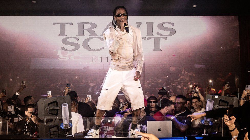 Travis Scott performing in Miami in July 2023. Travis, a black man with braids down to his shoulders, is on stage surrounded by a group of people holding phones and on laptops. He is wearing dark sunglasses, a beige coloured jacket, white shorts and white trainers.
