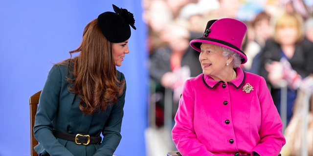 Kate Middleton in a hunter green dress and black fascinator chatting with Queen Elizabeth wearing a hot pink dress coat and matching hat