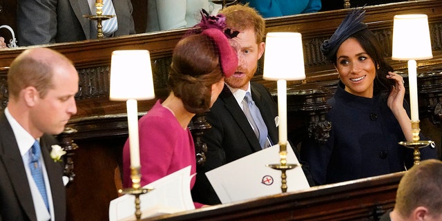 Kate Middleton wearing a pink dress and matching hat chatting with Prince Harry- in a suit and tie