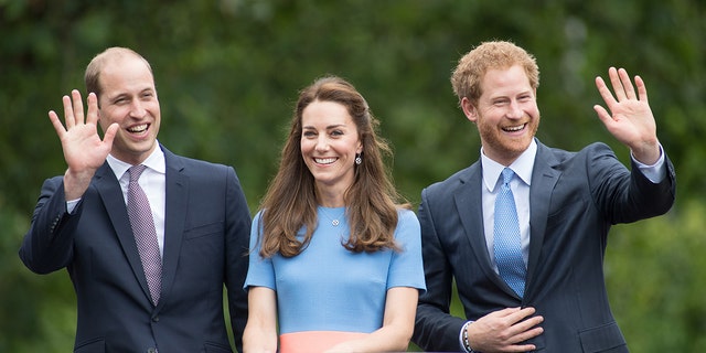 Kate Middleton wearng a light blue dress with a salmon-hued belt standing in between a smiling prince william and prince harry