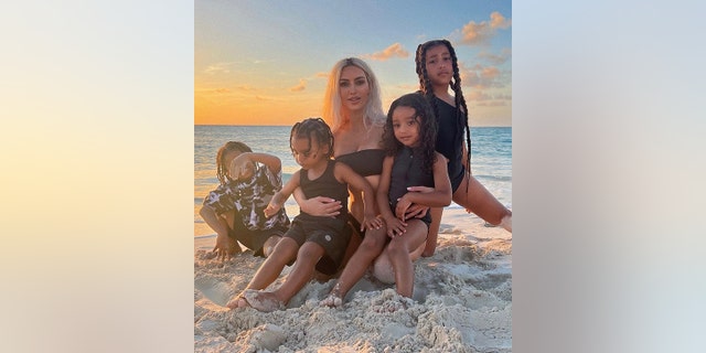 Kim Kardashian sits on the beach with her four children, balancing Chicago and Psalm on her laugh with North and Saint next to her