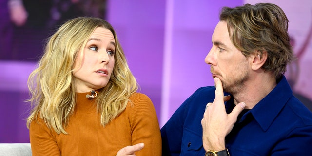 Kristen Bell in a burnt orange sweater looks towards Dax Shepard with a hand to his mouth while sitting on a couch during an interview